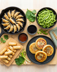 Vegan Asia SeriosFood - platou vegan asiatic pe blat bambus vedere de sus: gyoza vegetariene in tigaie fonta, mini spring rolls pe platou lemn, edamame, hotteok corean, sos de soia si sweet chili